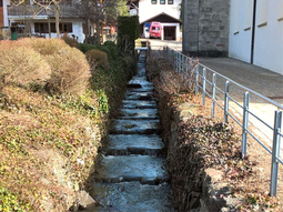 Bestehendes Gerinne am Burgberger Dorfbach entlang der Kirche. Auf den hier sichtbaren Abflussquerschnitt soll der Burgberger Dorfbach auch im Oberlauf verbreitert werden, um künftig hundertjährliche Hochwasserereignisse ohne Ausuferungen abführen zu können.